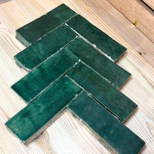 May include: A collection of rectangular ceramic tiles with a glossy, deep forest green finish, arranged in a classic herringbone pattern. Each tile features subtle variations in color depth and texture, showcasing a smooth, reflective surface.