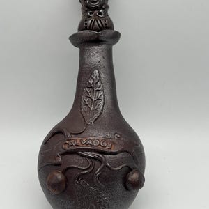 Könnte beinhalten: Eine dunkelbraune Keramikflasche mit einem dekorativen Verschluss, der ein lächelndes Gesicht mit Hut zeigt. Die Flasche hat eine strukturierte Oberfläche, geprägte Blatt- und Fruchtmotive und das Wort "CALVADOS" ist auf der Vorderseite geschrieben.