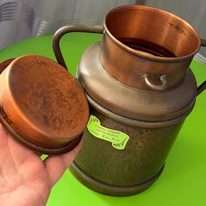 Op de afbeelding: Een koperen melkbus met een afneembaar deksel en twee handvatten aan de zijkant. De buitenkant heeft een donkere, verweerde patina, terwijl de binnenkant een heldere koperen afwerking heeft. Een lichtgroene sticker aan de zijkant bevat de tekst FABRICATION ARTISANALE Article Fait Main.