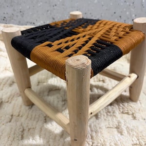 Könnte beinhalten: Kleiner, gewebter Hocker mit quadratischer Sitzfläche. Der Sitz ist schwarz und beige, die Beine sind aus hellem Holz. Der Hocker hat ein schlichtes, rustikales Design und eignet sich als Fußhocker oder dekoratives Element. Maße ca. 30 cm x 30 cm.