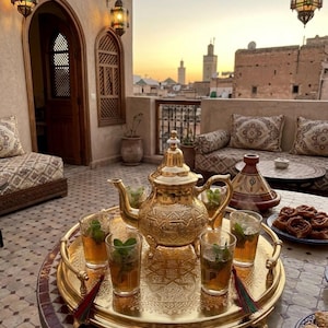 Peut inclure: Un service à thé doré, comprenant une théière et des verres remplis de thé et de menthe, est présenté sur un plateau décoratif. Le plateau repose sur une table à mosaïque, avec un coucher de soleil en arrière-plan.