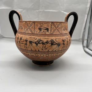 Könnte beinhalten: Eine terrakottafarbene Keramikschale mit zwei schwarzen Griffen. Die Schale ist mit schwarzen Figuren verziert, die Szenen von Tieren und Menschen darstellen. Der Rand weist ein geometrisches Muster auf. Diese Schale im antiken Stil ist ein dekoratives Stück.