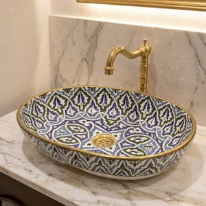 Peut inclure: Une vasque ovale en céramique ornée d'un motif géométrique bleu, vert et blanc, dotée d'un rebord doré et d'une bonde assortie. Un robinet doré haut et courbé est placé derrière la vasque, posée sur une surface en marbre blanc.