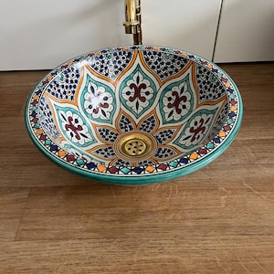 Może przedstawiać: Owalna ceramiczna umywalka ze szczegółowym wzorem kwiatowym w kolorze morskim, pomarańczowym i granatowym. Umywalka ma złoty kran i turkusową obwódkę. Umywalka jest umieszczona na drewnianej powierzchni.