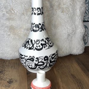 Könnte beinhalten: Eine weiße Keramikvase und ein passender kleinerer Verschluss, beide mit schwarzer arabischer Kalligrafie verziert. Die Vase hat einen hohen, schlanken Hals mit rotem Rand, während der kleinere Verschluss eine abgerundete Basis hat.