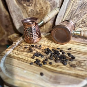 Könnte beinhalten: Zwei Kupfer-Kaffeekannen mit Holzgriffen liegen auf einem Holzschneidebrett, umgeben von Kaffeebohnen. Die Kannen haben eine gehämmerten Textur. Der Hintergrund zeigt weitere Holzbretter, was eine rustikale, warme Ästhetik erzeugt.