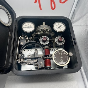 May include: A black industrial control box with the number 180 written in red above it. The interior contains three circular pressure gauges, two red wire coils, two maroon control knobs, and a pressure setting dial labeled PSI 0 to 200.