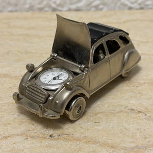 May include: A silver-toned metal miniature car figurine featuring an analogue clock face embedded in the bonnet. The car has a raised bonnet, a black roof section, and round headlights. The clock face is white with black numerals and hands.