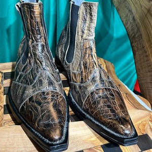 Könnte beinhalten: Ein Paar braune und schwarze Leder-Ankle-Boots mit Krokodilhaut-Textur. Die Stiefel haben eine spitze Zehenpartie und eine schwarze Sohle. Die Stiefel werden auf einer Holzoberfläche präsentiert.