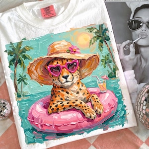 May include: White t-shirt featuring a cheetah wearing heart-shaped sunglasses and a sun hat, floating in a pink inner tube in a pool. The design includes palm trees, a cocktail, and a sunny beach scene.