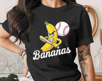May include: Black t-shirt with a cartoon banana character holding a baseball bat, and a baseball. The word "Bananas" is written in a script font. The design is a fun, playful graphic.