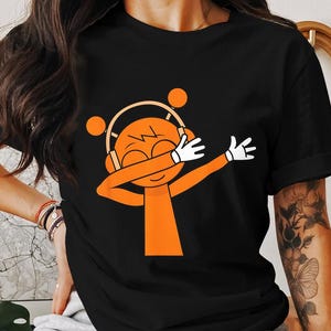 May include: Black t-shirt featuring an orange cartoon character wearing headphones and white gloves, with arms outstretched in a dabbing pose. The character has a smiling face and orange hair. The shirt is a casual wear item.