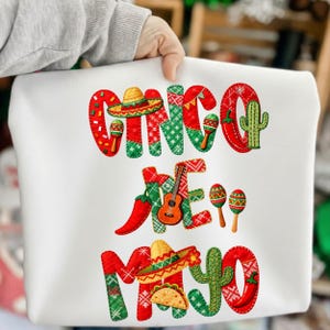 May include: White tote bag featuring the text "Cinco de Mayo" in red and green embroidery. The design includes a sombrero, maracas, a cactus, a guitar, a chili pepper, and a taco.