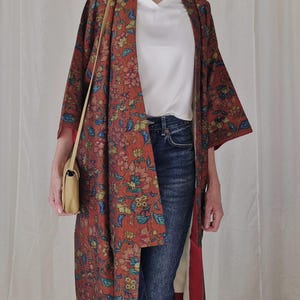 Pode incluir: Um longo quimono com estampa floral em tons de vermelho, azul e verde. O quimono está aberto, revelando uma blusa branca e jeans azuis. Uma pequena bolsa bege pende do ombro. A peça tem forro vermelho.