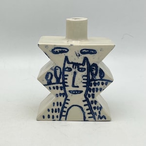 May include: A cream-colored ceramic vase with a geometric design. The vase features a blue cat-like face and abstract patterns. The vase has a cylindrical opening at the top and a stacked, zig-zag body. The vase is suitable for home decor.