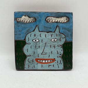 May include: A square ceramic tile featuring a whimsical monster design. The monster is light blue with a large smile and two small horns. The background has a blue sky with two white clouds and a green grassy area.