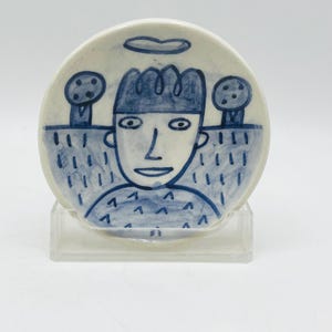 May include: A round ceramic plate featuring a hand-painted blue illustration of a person with a halo above their head, flanked by two stylized trees. The person has a textured shirt design and is set against a background of small blue marks.