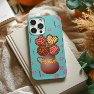 May include: A teal phone case featuring a colorful illustration of concha pastries in a brown ceramic mug. The design includes pink flowers and decorative elements. The phone case is resting on a beige book.