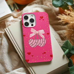 May include: A pink phone case featuring a cherry design with a pink bow and red hearts. The cherries have a volleyball pattern. The case is on top of a book and a beige blanket.