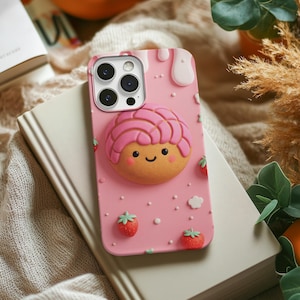 May include: Pink phone case featuring a 3D concha design with a smiling face and pink frosting. The case also has strawberry and cloud accents. The phone case is resting on a book.