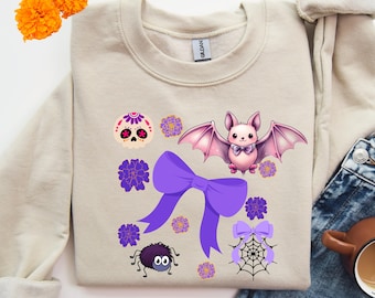 May include: Beige sweatshirt featuring Halloween-themed graphics. The design includes a purple bow, a cute bat with pink wings, a spider, a sugar skull, and floral accents. The sweatshirt is a cozy, casual garment.