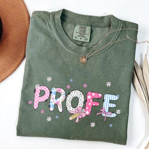 May include: Olive green Comfort Colors t-shirt with the word "PROFE" in a colorful, patterned design. The letters are decorated with pink stars, blue flowers, and a pencil with a pink bow. A gold necklace with a pendant is draped across the shirt.