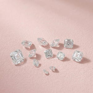 May include: A collection of clear, faceted diamonds of various shapes and sizes, scattered on a soft pink surface. The diamonds include emerald cuts and other geometric forms, reflecting light and creating a sparkling effect.