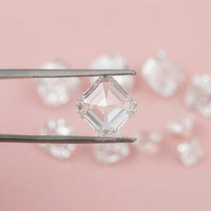 May include: A clear, square-cut gemstone is held by tweezers against a soft pink background. The gemstone has multiple facets, reflecting light. Other out-of-focus gemstones are visible in the background. The text "calijewels" is visible on the tweezers.