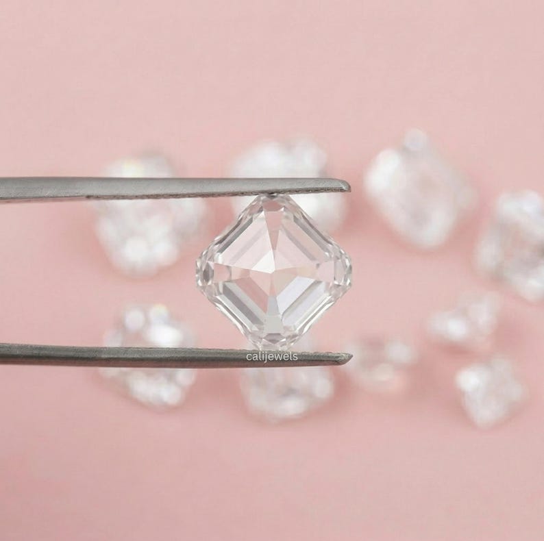 May include: A clear, square-cut gemstone is held by tweezers against a soft pink background. The gemstone has multiple facets, reflecting light. Other out-of-focus gemstones are visible in the background. The text "calijewels" is visible on the tweezers.