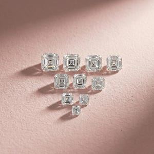 May include: An assortment of clear, square-cut gemstones of varying sizes, arranged on a pink surface. The gemstones are faceted, reflecting light and displaying internal patterns. The brand name "calijewels" is visible.