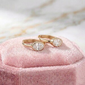 May include: A pair of gold hoop earrings with oval-cut, clear gemstones. The earrings are displayed on a pink velvet surface, with a blurred marble background. The earrings are a classic jewelry item.