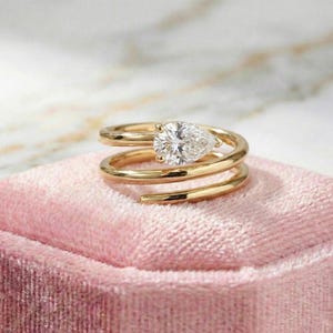 May include: A gold ring with a pear-shaped diamond set in a spiral design. The ring is displayed on a pink velvet surface, showcasing its intricate details and elegant style. The ring's band wraps around the finger in a unique, modern design.