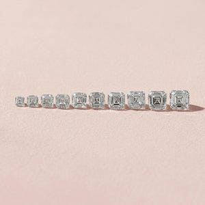 May include: A collection of Asscher-cut diamond gemstones of varying sizes, arranged in a row on a light pink surface. The diamonds are clear and reflect light, showcasing their square shape with cut corners and step-cut facets.