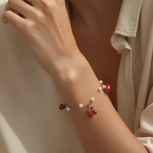 Op de afbeelding: Een gouden armband met rode kersen- en aardbeienbedels, geaccentueerd met kleine witte parels. Een gouden ring met een spiraalvormig ontwerp is ook zichtbaar. De sieraden worden gedragen om de pols en vinger van een persoon.