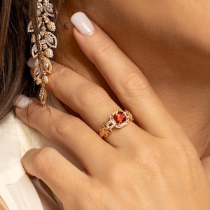 Peut inclure: Une bague en or avec une pierre précieuse rouge entourée de petites pierres précieuses claires. La bague est portée au doigt d'une personne.