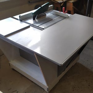 May include: A white table saw with a green and black blade, mounted on a white wooden base with a shelf. The saw has a red lever and a white extension table.