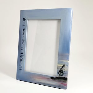 May include: A light blue wooden picture frame with a painted landscape scene. The frame has the words "Memories are made here" printed on the side.