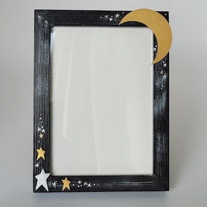 May include: Black picture frame with a white distressed finish. The frame has a gold crescent moon and stars painted on the top right corner and gold and white stars painted on the bottom left corner.