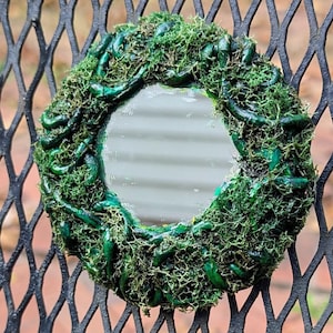 May include: A small, round mirror is surrounded by a wreath of green moss and decorative green accents. The mirror is mounted on a black metal fence with a diamond pattern. Decorative metal flowers are at the top of the fence.