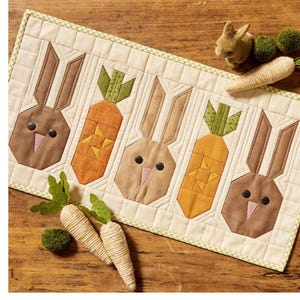 May include: A quilted Easter table runner featuring five geometric shapes: three brown bunnies and two orange carrots with green tops. The runner is bordered with a green and white checkered pattern. Decorative carrots and a small bunny are also present.