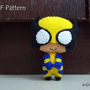 Puede incluir: Un juguete de fieltro amarillo con una máscara de fieltro negro y ropa de fieltro azul. El juguete tiene una cara de fieltro blanco con ojos negros y una boca sonriente. El juguete está etiquetado como "PDF Pattern" en la parte superior de la imagen.