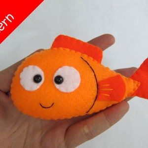 May include: A felt orange fish with black eyes, a white mouth, and a red tail fin. The fish is being held in a hand.