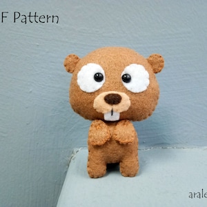 May include: A brown felt stuffed animal beaver with white felt eyes and a brown felt nose. The beaver is standing on a light blue surface. The text "PDF Pattern" is at the top of the image and the text "araleing" is at the bottom of the image.