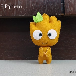 Puede incluir: Un juguete de fieltro de un personaje con un cuerpo marrón, ojos blancos y una boca negra. El personaje tiene dos hojas verdes en la parte superior de su cabeza. El juguete está de pie sobre una superficie marrón. El texto "PDF Pattern" es visible en la parte superior de la imagen.