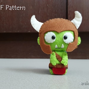May include: A green felt plush toy with a brown helmet and white horns. The toy has a friendly face with large eyes and a small mouth. The toy is wearing a red loincloth. The image includes the text "PDF Pattern".