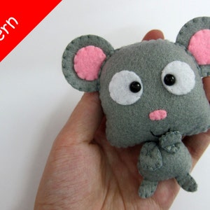 May include: A gray felt mouse with pink ears and white eyes. The mouse is holding its paws together in front of its chest. The mouse is being held in a person's hand.