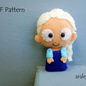 May include: A handmade felt doll of a young girl with long white hair, wearing a blue dress and light blue sleeves. The doll has a smiling face with large brown eyes and white eyelids. The doll is standing on a white surface with a blue wall in the background. The text "PDF Pattern" is visible in the top left corner of the image.