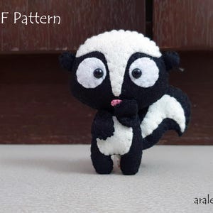 May include: A black and white plush toy skunk with a pink nose and black eyes. The toy is standing on a white surface.  PDF Pattern