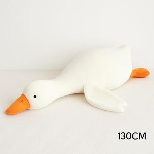 May include: A plush toy shaped like a white goose with an orange beak and orange feet. The goose is in a lying position with its head extended forward. The text 130CM is printed in black in the lower right corner of the image.
