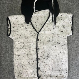 May include: A hand-knitted baby sweater and hood set. The sweater is speckled white with black flecks, a V-neck, and black trim. The hood is black with a white trim. The sweater has black buttons.